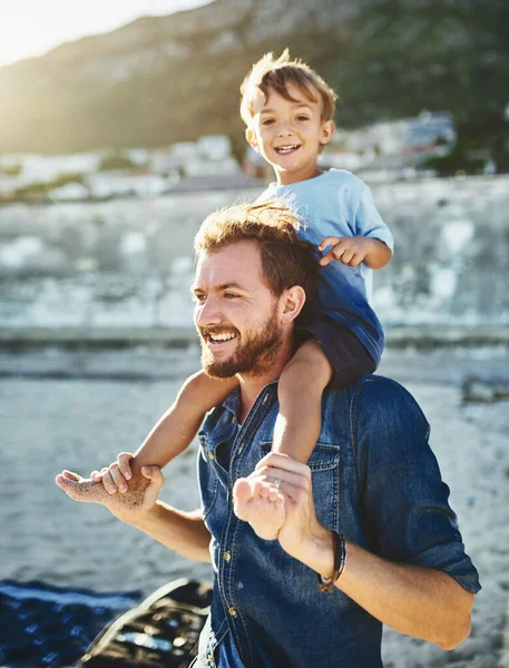 Sitting on Daddys shoulders makes me feel taller. Shot of a young Father and son spending quality time at the beach. — Foto de Stock