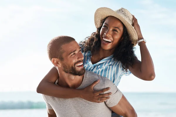 Summer keeps them smiling. Shot of a young man piggybacking his girlfriend at the beach. — ストック写真