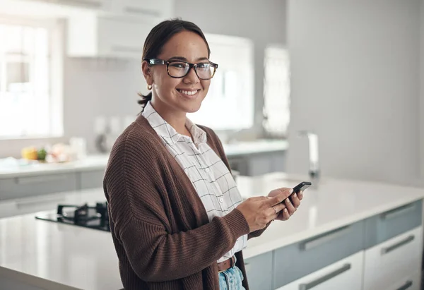 Just checking up on some notes on my phone. Portrait of a young woman using a cellphone in the kitchen at home. — 스톡 사진