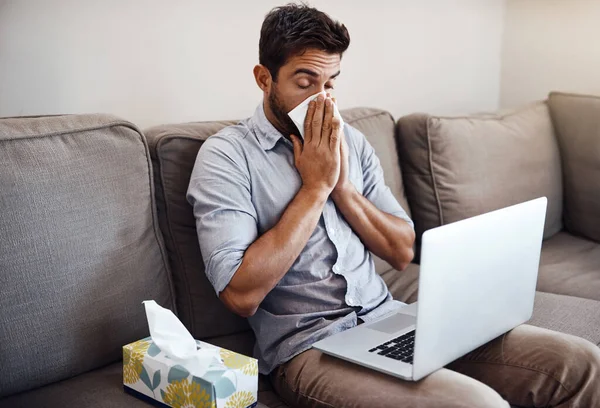 Hopefully he can mange to finish all his work today. Shot of a young businessman blowing his nose with a tissue while trying to work on his laptop at home. — Stock Photo, Image