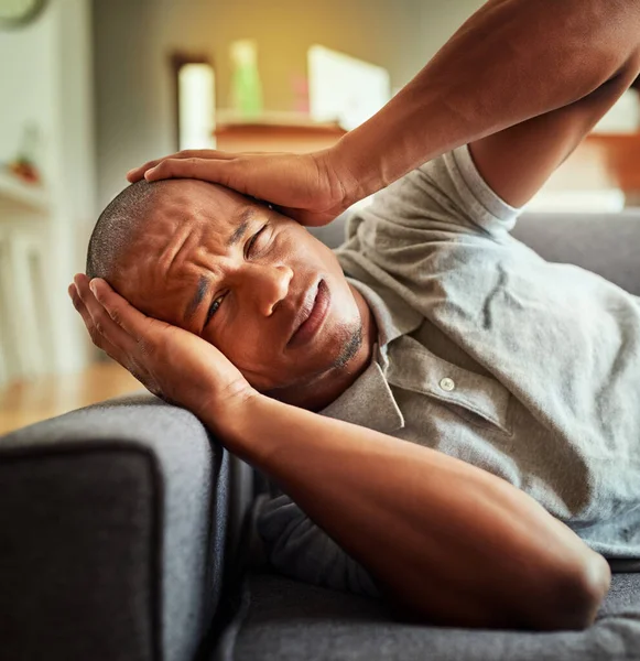 My brain cant take it anymore. Shot of a stressed out young man lying on a couch while holding his head in discomfort inside at home during the day. — Stock Photo, Image