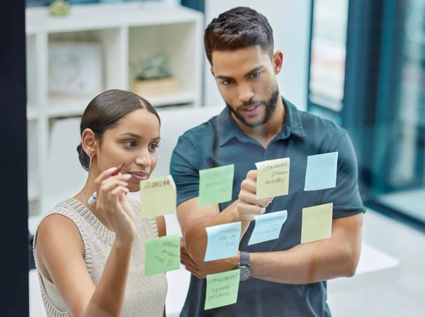 Lets figure this out.... Shot of two businesspeople brainstorming with sticky notes on a transparent wall. — Stock Photo, Image