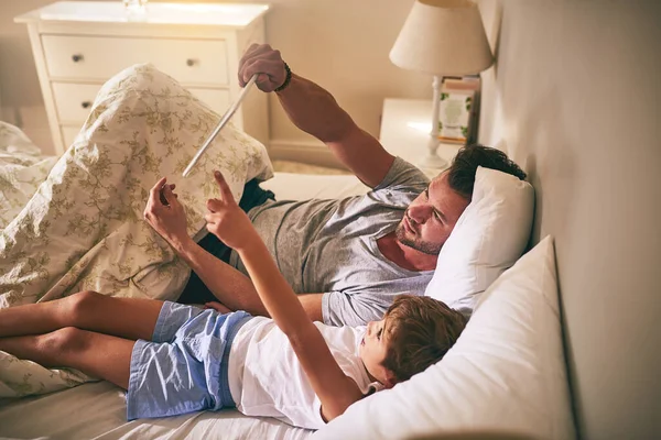 Laten we daarnaar kijken. Schot van een vader en zijn zoontje met behulp van een digitale tablet terwijl ze thuis in bed liggen. — Stockfoto