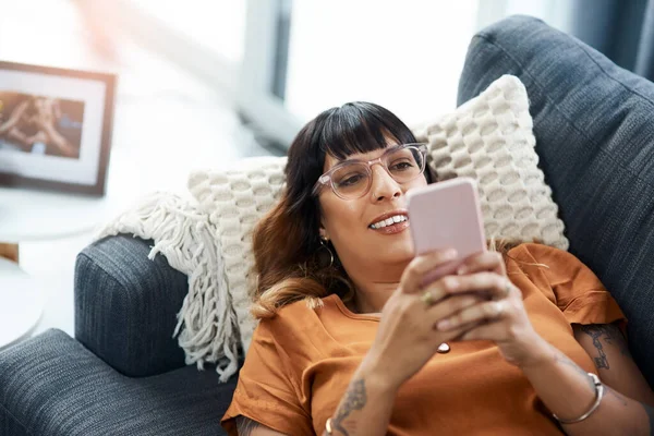 Escolhendo um dos muitos aplicativos de mídia social para matar o tédio. Tiro cortado de uma jovem mulher usando seu celular enquanto relaxa no sofá em casa. — Fotografia de Stock