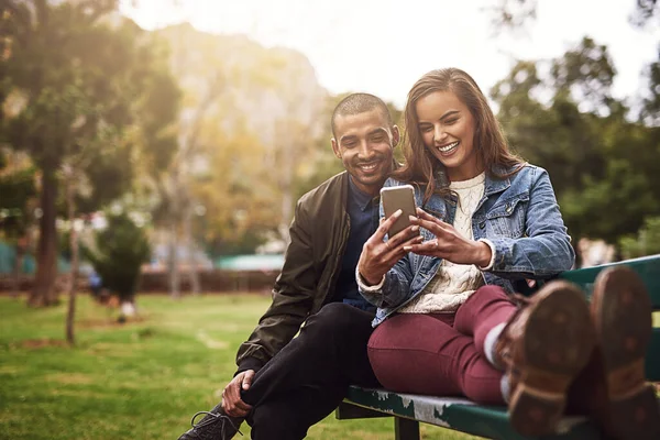 ¿Qué opinas de esta foto? Toma de una alegre pareja joven sentada en un banco mientras usan un teléfono juntos afuera en un parque. —  Fotos de Stock