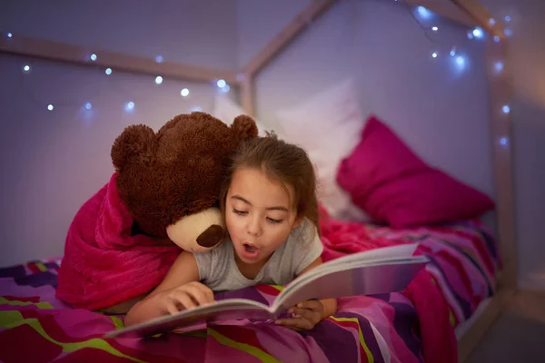 To opowieść pełna wspaniałych niespodzianek. Przycięte ujęcie małej dziewczynki czytającej książkę w łóżku ze swoim misiaczkiem. — Zdjęcie stockowe