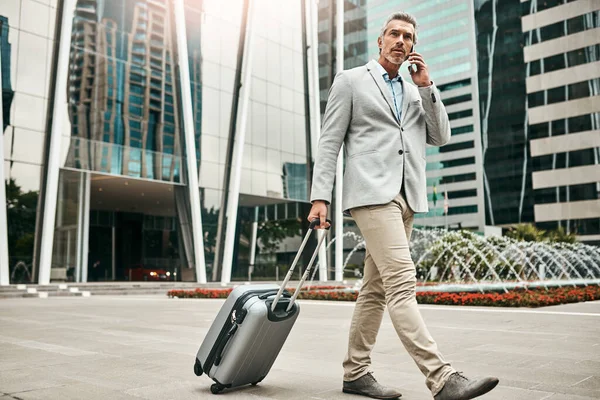 Sono atterrato e sono pronto a parlare di affari. Girato di un uomo d'affari maturo che parla al cellulare mentre cammina con una valigia in città. — Foto Stock