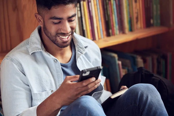 Se alguém precisar de mim, estarei com os livros. Tiro de um jovem usando um smartphone em uma biblioteca na universidade. — Fotografia de Stock