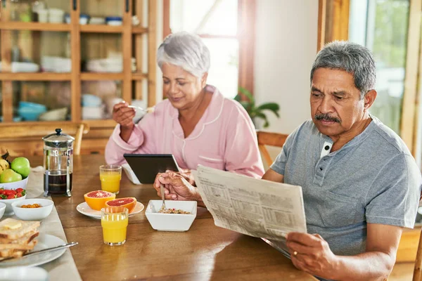 Читання новин за допомогою двох різних методів. Знімок безтурботної пари літніх людей, яка поснідала разом, читаючи папір і переглядаючи цифровий планшет вдома . — стокове фото