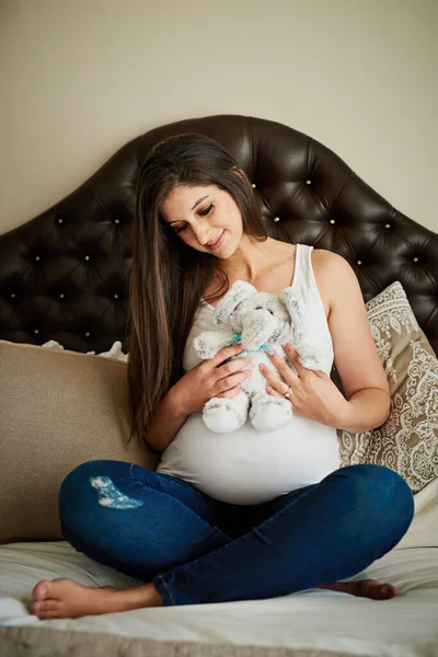Non vedo l'ora di giocare con te. Girato di una donna incinta felice in possesso di un giocattolo di peluche mentre seduto sul suo letto a casa. — Foto Stock