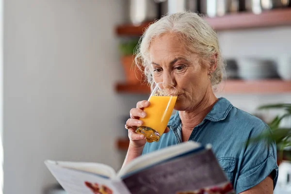 Não é uma dieta, é um modo de vida. Tiro de uma mulher madura lendo um livro enquanto toma suco de laranja em casa. — Fotografia de Stock