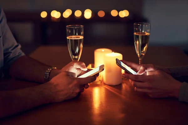 Dejemos que todos sepan que estaban comprometidos. Cortado de una pareja irreconocible mensajes de texto en sus teléfonos móviles durante una cena encendida con velas por la noche. — Foto de Stock
