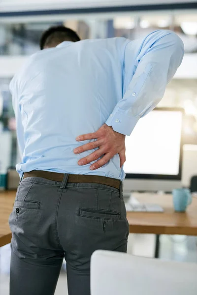 Hes been sitting down for too long. Rearview shot of a young businessman holding his back while standing at his desk. — Stock Photo, Image