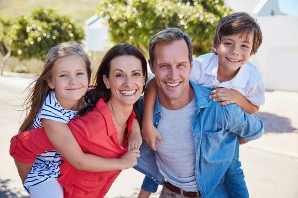 Draußen etwas Familienspaß genießen. Porträt lächelnder Eltern, die ihrem kleinen Sohn und ihrer Tochter an einem sonnigen Tag huckepack draußen auf ihrem Hof geben. — Stockfoto