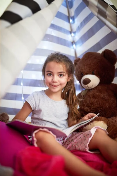 子供は子供になる。女の子の肖像画読書A本とともに彼女のteddybearでAテントの中で自宅. — ストック写真