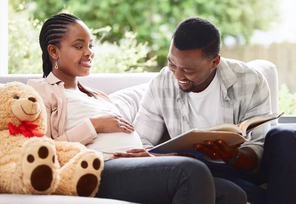 He loves reading to his unborn baby. Cropped shot of a handsome young man reading a book to pregnant wifes belly while sitting on the sofa at home.