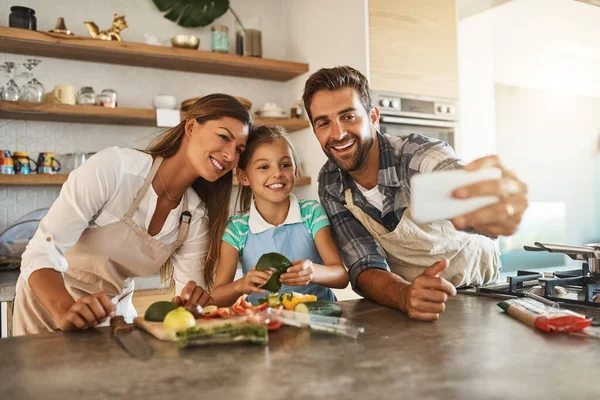 Ehhez kell egy szelfi. Egy boldog fiatal család fotózása, akik egy szelfihez pózolnak miközben együtt főznek a konyhájukban.. — Stock Fotó