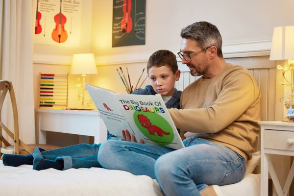 Μαθαίνοντας για την ιστορία αυτών των συναρπαστικών πλασμάτων. Πυροβόλησε έναν πατέρα και ένα γιο διαβάζοντας ένα βιβλίο για δεινόσαυρους μαζί σε ένα υπνοδωμάτιο στο σπίτι. — Φωτογραφία Αρχείου