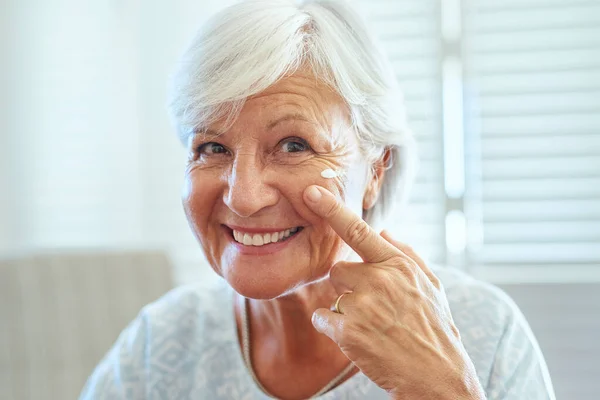 现在是时候照顾你的皮肤了。一位快乐的老年妇女在家里用护肤霜贴脸的画像. — 图库照片