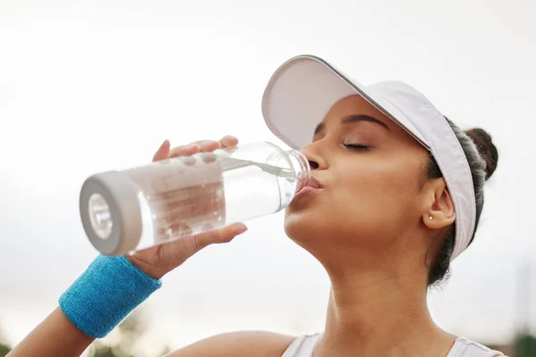 Není lepší způsob, jak uhasit žízeň. Shot krásné mladé ženy pití láhev vody, zatímco hraje tenis. — Stock fotografie