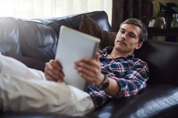Surfen op zijn bank. Hoge hoek foto van een knappe jonge man met behulp van zijn tablet terwijl zitten op de bank thuis. — Stockfoto