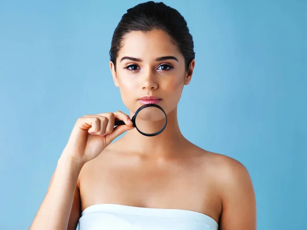 Hautpflegegewohnheiten in den Fokus rücken. Studioporträt einer schönen jungen Frau mit einer Lupe vor blauem Hintergrund. — Stockfoto