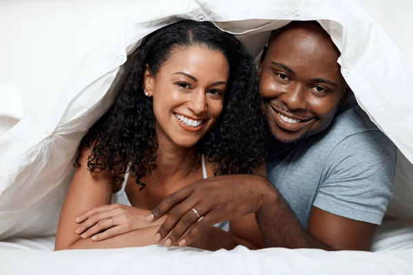 Alltid på gott humör tillsammans. Porträtt av ett muntert ungt par som ligger i sängen tillsammans under en filt medan du tittar på kameran hemma på morgonen. — Stockfoto