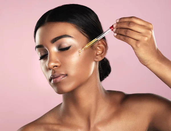 Su piel será la envidia de todos. Foto de estudio de una hermosa joven que se aplica aceite esencial en la cara con un gotero posando sobre un fondo rosa. — Foto de Stock