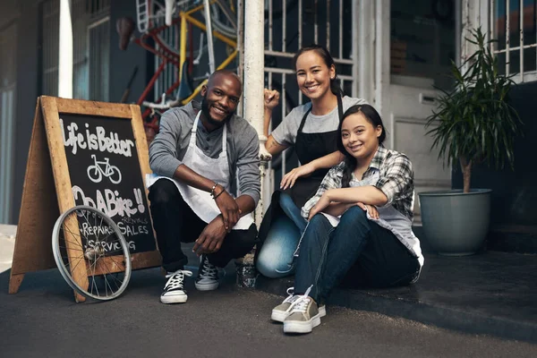 Tenemos lo que tú quieres. Retrato de tres jóvenes trabajadores sentados fuera de su taller de reparación de bicicletas. — Foto de Stock