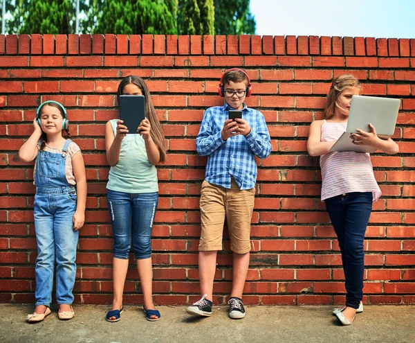 La infancia en la era digital. Fotografía de un grupo de niños pequeños usando dispositivos digitales contra una pared de ladrillo. —  Fotos de Stock