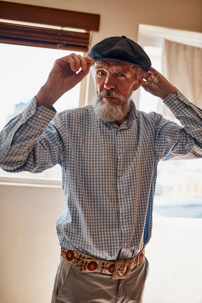 Er ist ein stilvoller Senior. Porträt eines fröhlichen älteren Mannes, der einen seiner Hüte anprobiert, während er in die Kamera blickt. — Stockfoto