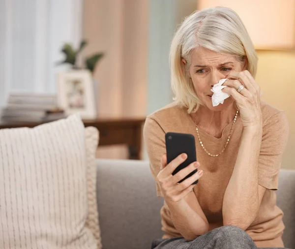 2020, the year marked by trauma. Shot of a mature woman using a smartphone and crying on the sofa at home. — Stock Photo, Image