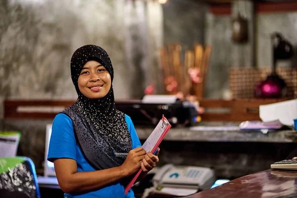 Siete sempre i benvenuti qui. Ritratto di una donna thai musulmana sorridente in piedi dietro una reception di un hotel con una cartellina. — Foto Stock