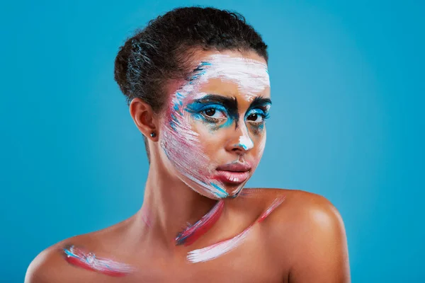 Brilhante, arrojado e bonito. Estúdio retrato de uma bela jovem coberta de pintura facial posando contra um fundo azul. — Fotografia de Stock