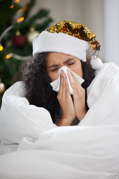 Shes got a case of the Christmas sniffles. Cropped shot of an attractive young woman blowing her nose while wearing a Christmas hat and lying on the sofa at home. — Stock Photo, Image