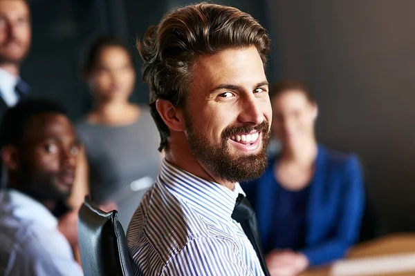 Ele lidera sua equipe através da positividade. Retrato de um empresário sorridente sentado em um escritório com colegas em segundo plano. — Fotografia de Stock