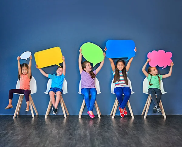 Llena el mundo con tu voz. Foto de estudio de un grupo diverso de niños sentados en sillas y sosteniendo burbujas de habla sobre un fondo azul. —  Fotos de Stock