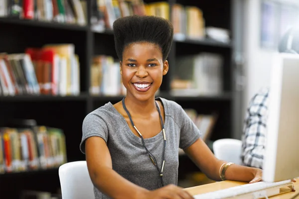 Örülök, hogy dolgozhatok a jövőmön. Egy vidám fiatal nőről, aki számítógépen dolgozik a könyvtárban, miközben a kamerába néz.. — Stock Fotó