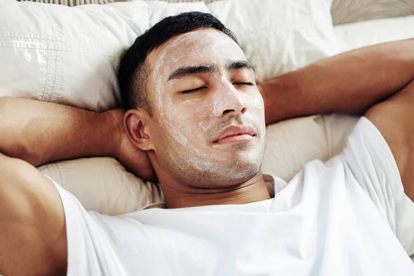 Take a some time out for yourself, you deserve it. Shot of a man wearing a cosmetic face mask while lying on his bed at home. — Stock Photo, Image