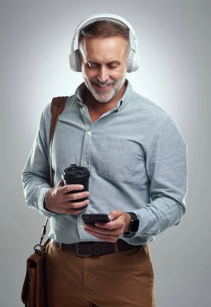 Es un buscavidas que siempre está preparado para el día. Foto de estudio de un hombre maduro que usa auriculares y un teléfono celular mientras lleva una bolsa y una taza de café sobre un fondo gris. — Foto de Stock