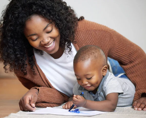 What masterpiece are you creating now. Shot of an attractive young mother lying on the living room floor with her son and watching him draw.