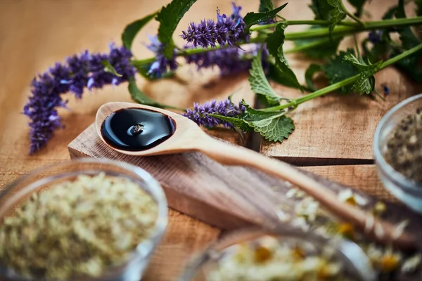 Há muito que se pode fazer com a lavanda. Tiro de uma colher de madeira com o líquido escuro nele rodeado por lavanda fresca em uma mesa. — Fotografia de Stock