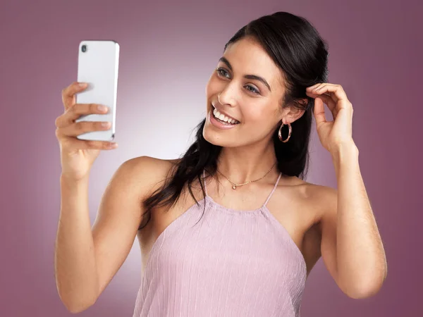 Look for the magic in every moment. Shot of a beautiful young woman holding up her mobile phone to take a selfie while adjusting her hair behind her ear against a pink background. — Stock Photo, Image