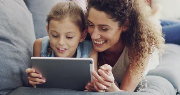 Делится своими дочерьми интернет-интересами. 4k видеозапись матери и ее маленькой дочери, лежащей вместе на диване дома с помощью цифрового планшета. — стоковое видео