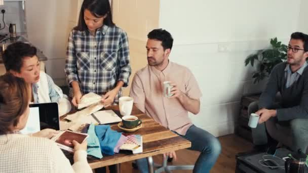 Διαφορετική ομάδα επιχειρηματιών που κοιτάζουν φωτογραφίες και πίνουν καφέ κατά τη διάρκεια μιας συνάντησης. Μια νεαρή επιχειρηματίας που κοιτάζει δείγματα υφάσματος ενώ οι συνάδελφοί της κάνουν brainstorm και κοιτάζουν φωτογραφίες — Αρχείο Βίντεο