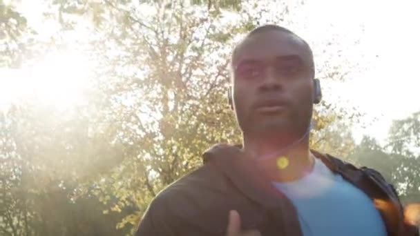 Focused young african american jogging through a park and listening to music on his headphones. Serious young man working out and running through a garden in slow motion listening to music. — Wideo stockowe