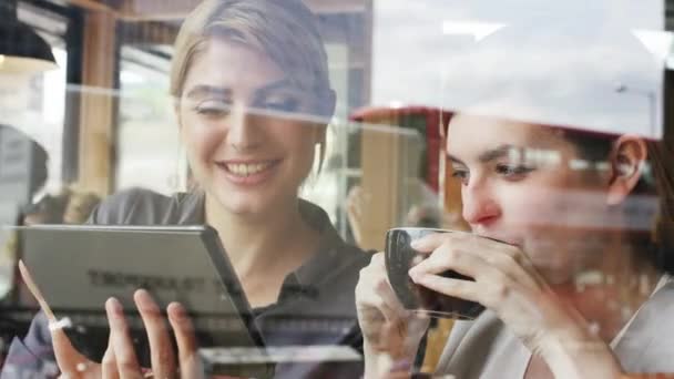 Two businesswomen planning during a meeting in a cafe while drinking coffee and using a wireless tablet. Two smiling businesswomen collaborate and talk while scrolling online on a digital tablet. — Vídeo de stock