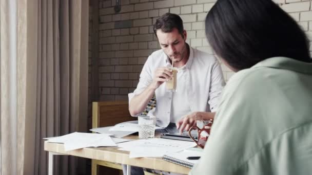 A young businessman drinking coffee during a meeting looking over architectural drawings with colleagues in a cafe. Diverse group of businesspeople collaborating and brainstorming reading documents — стоковое видео