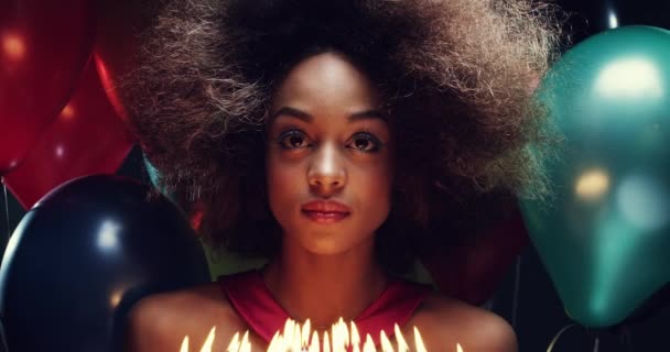 Jovem mulher em sua festa de aniversário comemorando, segurando um bolo com velas. Jovem afro-americana com um afro segurando seu bolo de aniversário olhando para as velas cercadas por balões coloridos — Vídeo de Stock