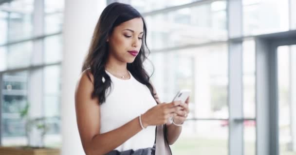 De zaken gaan maar door, hou het tempo bij. 4k videobeelden van een jonge zakenvrouw met behulp van een smartphone tijdens het lopen door een modern kantoor. — Stockvideo